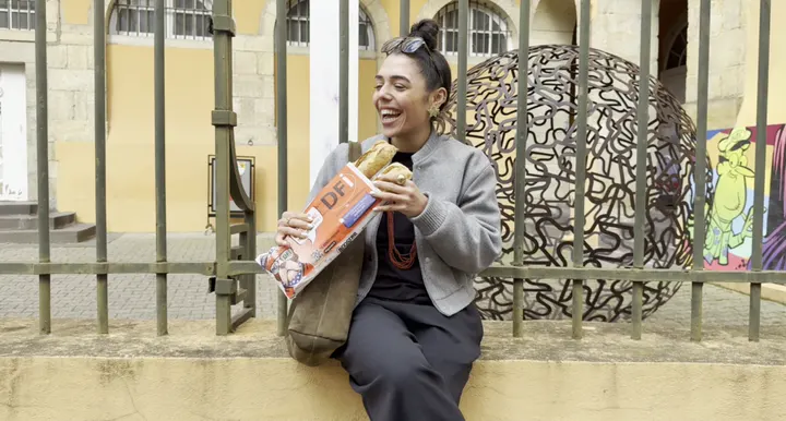 Jeune femme qui croque une baguette dans son sac à pain publicitaire, assise devant une grille : tournage Booster Media pour les réseaux sociaux